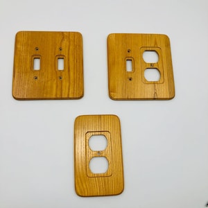 May include: Three light brown wooden wall plates with a visible grain pattern. One plate features two toggle switch openings, one plate has a single toggle switch and a duplex outlet opening, and one plate has a single duplex outlet opening.