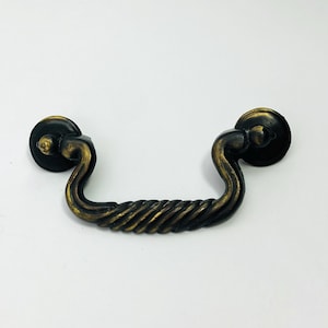 May include: Antique-style drawer pull with a dark bronze finish. The handle has a twisted rope design in the center, with circular mounting plates on each end. The hardware is designed for cabinets and drawers.