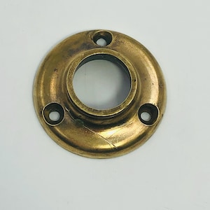 Può includere: Un elemento circolare in ottone con un foro centrale e tre fori più piccoli distribuiti uniformemente sul perimetro. L'ottone ha un aspetto leggermente invecchiato e ossidato, suggerendo che sia vintage o antico. È probabilmente un elemento decorativo o funzionale.