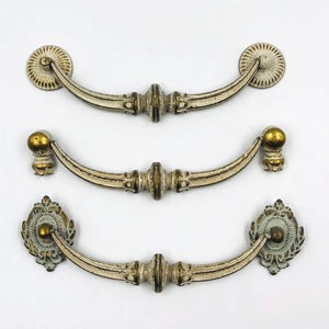 May include: Three antique-style drawer pulls with a distressed white finish and gold accents. The pulls have a curved design with decorative details and are suitable for cabinets or furniture. Each pull measures approximately 15 cm in length.
