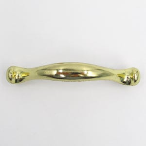 May include: A gold-coloured cabinet pull handle. The handle has a curved, elongated design with decorative detailing at each end. The hardware is made of metal and is set against a white background.
