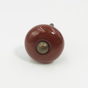 May include: A round, glossy, dark reddish-brown ceramic cabinet knob with a smooth, reflective surface. The center of the knob features a small, circular, metallic-toned hardware component that is slightly recessed into the ceramic body.