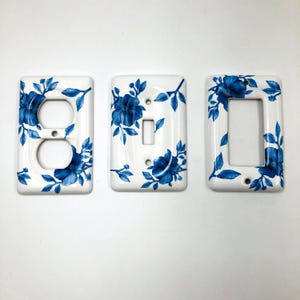 Könnte beinhalten: Drei weiße, rechteckige Wandabdeckungen mit blauem Blumenmuster. Das Set besteht aus einer Abdeckung für eine Doppelsteckdose, einer für einen Kippschalter und einer für einen rechteckigen Wippschalter, alle mit blauen Blüten.