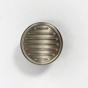 May include: A circular cabinet knob with a brushed metallic finish. The face of the knob features a series of horizontal, raised parallel lines that span across the center, creating a textured, linear pattern within the round frame.