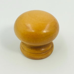 Puede incluir: Un pomo de madera para armario con una parte superior redondeada y una base circular. El pomo está hecho de madera de color claro y tiene un acabado liso y pulido. La base presenta dos anillos concéntricos. Este herraje es adecuado para muebles.