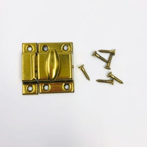 May include: A brass-colored metal cabinet latch featuring a rectangular base with a central oval-shaped rotating knob and four mounting holes. The hardware set includes four matching brass-colored screws with threaded shanks and flat heads.