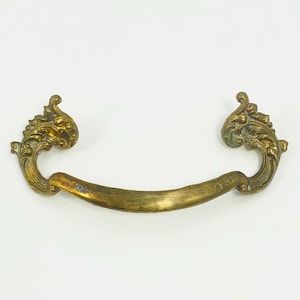 May include: Ornate brass drawer pull with a curved handle and decorative, scroll-like ends. The brass has a weathered, antique appearance, with variations in color from gold to a darker, tarnished hue. This hardware is suitable for vintage furniture restoration.