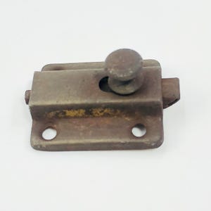 May include: A vintage, rusty metal latch with a circular knob. The latch has a rectangular base with two holes and a small, protruding piece on the side. The metal has a weathered appearance.