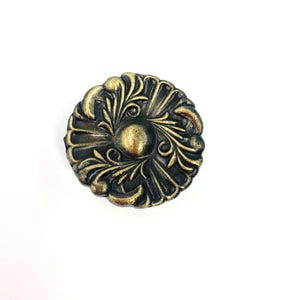 Puede incluir: Un pomo de mueble redondo y decorativo con un diseño floral ornamentado. El pomo presenta una base de bronce oscuro con detalles dorados, creando una estética vintage. El elemento central del diseño es un medallón circular en relieve.