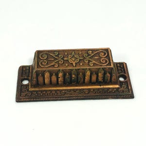 Puede incluir: Tirador de cajón ornamental de color bronce con intrincados diseños florales y de volutas. El mango rectangular presenta un panel central elevado con un patrón decorativo y dos orificios de montaje en cada extremo. Una pieza de ferretería de estilo vintage.
