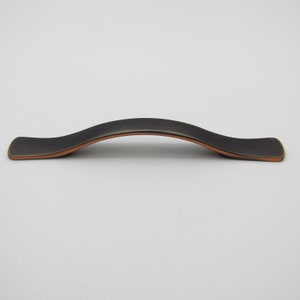 Puede incluir: Un tirador de armario negro con un acento de cobre. El tirador es curvo y tiene una forma rectangular plana.