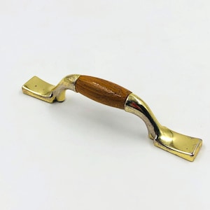 Könnte beinhalten: Ein Schrankgriff mit einem Mittelteil aus Holz und goldfarbenen Metallenden. Der Griff hat ein gebogenes Design und ist vor einem weißen Hintergrund platziert. Der Holzabschnitt hat eine natürliche Maserung.