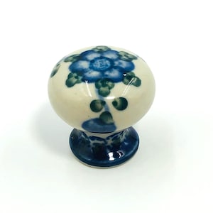Könnte beinhalten: Ein Keramik-Schrankknopf mit floralem Design. Der Knopf ist hauptsächlich cremefarben mit einem blauen Blumenmuster auf der Oberseite und einem blauen Sockel. Das florale Design umfasst blaue und grüne Akzente. Der Knopf ist rund und hat eine Pilzform.