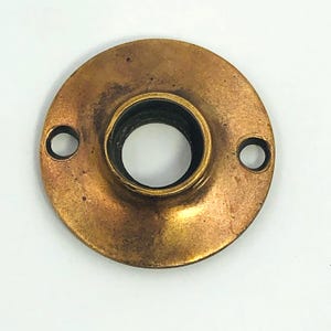 以下が含まれることがあります： 円形のデザインのヴィンテージ真鍮製ドアノブバックプレート。プレートには中央の穴とネジ用の2つの小さな穴があります。真鍮は暖かく、古くなった風合いで、年齢と使用を暗示する暗い部分があります。 全体的な形状は丸いです。