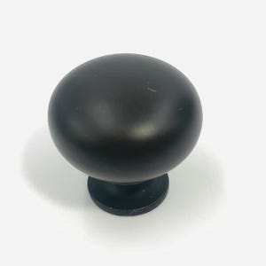 May include: A matte black cabinet knob featuring a smooth, rounded, mushroom-shaped head that tapers down to a circular base. The hardware has a solid, uniform dark finish with a clean, curved silhouette designed for attachment to furniture.