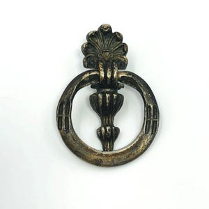 May include: Antique silver-toned drawer pull with a decorative design. The pull features a circular handle with a central, ornate floral-like element and a decorative top. The metal has a tarnished appearance, suggesting age.