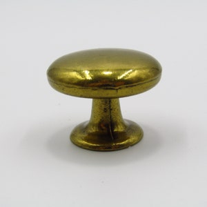 Puede incluir: Un pomo de armario en forma de hongo de color dorado, con una parte superior redonda y lisa y una base ligeramente acampanada.