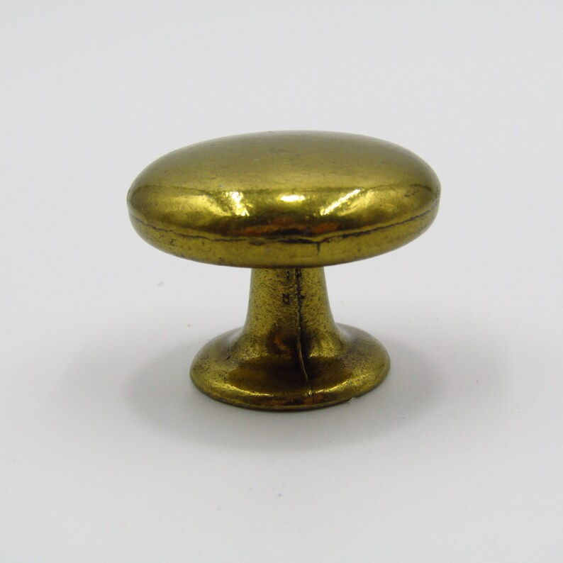 Puede incluir: Un pomo de armario en forma de hongo de color dorado, con una parte superior redonda y lisa y una base ligeramente acampanada.