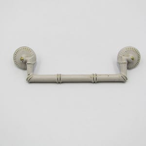 Puede incluir: Un tirador de armario gris claro con un diseño de estilo bambú. El tirador tiene dos piezas finales circulares con un patrón en espiral y una barra de conexión con detalles de bambú. Se ven pequeños detalles de color latón.