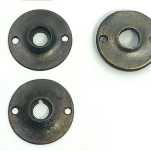 以下が含まれることがあります： アンティークブロンズ色の金属製ドアハードウェアのロゼット3つ。各ロゼットは円形で、中央に穴が1つ、取り付け用の小さな穴が2つあります。金属は風化し、古びた外観をしています。