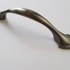 Puede incluir: Un tirador de armario curvo, gris oscuro, con acabado metálico cepillado. El tirador tiene un diseño suave y ergonómico y está montado sobre dos bases decorativas y redondeadas. Ideal para armarios de cocina o baño.