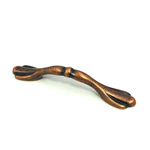May include: A bronze-colored cabinet pull with a curved design. The handle features a central, slightly raised section and flared ends. The finish has a slightly textured appearance, adding visual interest to the hardware.