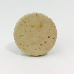 Puede incluir: Un pomo de cerámica circular con un acabado liso y brillante en color beige claro. La superficie presenta un patrón sutilmente moteado con pequeñas manchas marrones y tostadas dispersas, creando un aspecto natural y texturizado.