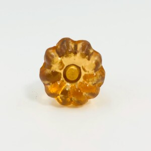 May include: A translucent amber-colored glass knob with a scalloped, flower-like edge and a circular center indentation. The object features a glossy, reflective surface with a warm, golden-brown hue and a distinct, multi-petaled radial design.
