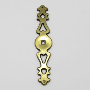 Pode incluir: Puxador de armário decorativo em tom de latão. O puxador apresenta recortes em forma de coração, um elemento circular central com um orifício quadrado e extremidades decorativas. A ferragem é projetada para móveis.