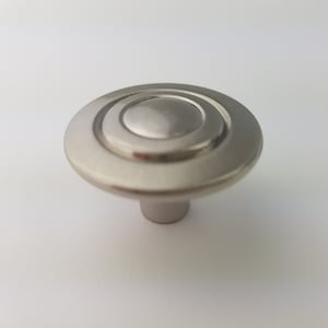 May include: A silver cabinet knob with a round, slightly raised center and a concentric circle design.