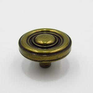 May include: Antique brass cabinet knob with a circular design. The knob features concentric rings and a central, raised circle. The finish has a vintage, aged appearance, suggesting a classic or retro style. The knob is designed for easy installation.