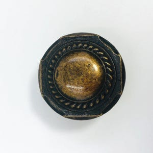 May include: A round cabinet knob with a dark, weathered bronze finish. The center features a smooth, raised dome surrounded by a textured rope-style border. The outer edge has a slightly octagonal shape with a distressed, dark patina finish.