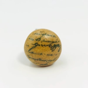 May include: A round, beige cabinet knob with a smooth, speckled finish. The surface is decorated with black, cursive handwritten-style text that wraps around the sphere, featuring legible script and ink-like markings across the entire front.