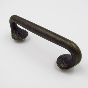 May include: A dark brown, hammered metal cabinet pull with a curved design. The pull has a textured finish and a slightly rounded shape.