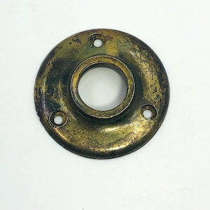May include: A vintage brass door knob backplate with a circular design. The backplate has a central hole and three smaller holes evenly spaced around the perimeter. The brass has a tarnished, aged appearance.