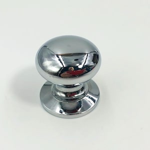 May include: A polished chrome cabinet knob featuring a smooth, rounded mushroom-shaped head and a circular base plate. The metallic surface is highly reflective, showing a clear mirror-like finish that captures the surrounding light and environment.