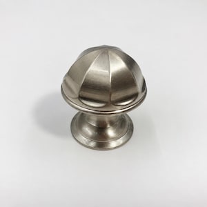 May include: A brushed nickel cabinet knob featuring a rounded, dome-shaped top with vertical fluted ridges. The hardware has a circular base that tapers upward to support the textured, segmented head, providing a metallic finish throughout.