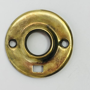 May include: A circular, brass-colored door plate with a central opening and two smaller holes. The plate has a rectangular cutout at the bottom and shows signs of wear, suggesting age or use. The hardware is likely for a door handle or lock.