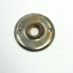 May include: A circular, vintage metal door knob backplate with a tarnished silver finish and a brass-coloured centre. Two small holes are visible on the plate. The backplate is likely for a door or cabinet.
