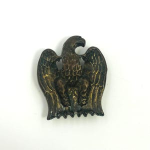 May include: A metallic eagle emblem with a dark bronze finish, featuring detailed feather textures on the wings and body. The eagle is depicted in a standing position with its head turned to the right, showcasing a weathered, aged patina.