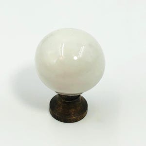 May include: A spherical white knob with a smooth, polished finish attached to a circular, dark brown, textured metal base. The object features a rounded top section that sits securely on the darker, metallic mounting hardware component.