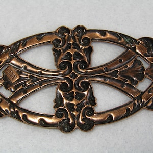 May include: Ornate copper-colored metal decorative piece with an openwork design. The piece features a central floral motif and scrolling details. The text "GENUINE COPPER" is visible. The overall shape is oval, with a small hole at one end.