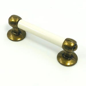 Pode incluir: Um puxador de armário com um centro de cerâmica branca e extremidades de cor latão antigo. O puxador tem uma forma cilíndrica com extremidades arredondadas e bases circulares. O hardware é projetado para móveis e armários.