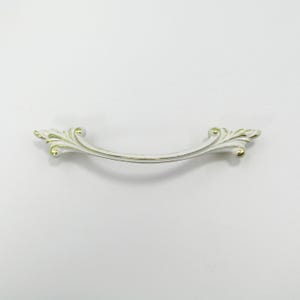 Könnte beinhalten: Dekorativer Schubladengriff in Weiß und Gold mit gebogenem Griff. Die Enden weisen verzierte, blattartige Designs auf. Der Griff ist etwa 15 cm lang.