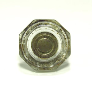 Pode incluir: Puxador de vidro transparente e facetado com centro metálico. O puxador tem formato octogonal e estética vintage. O centro metálico é de cor prata fosca. O vidro é transparente.