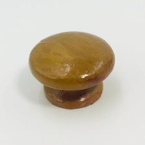 May include: A round, mushroom-shaped cabinet knob with a smooth, glossy finish in shades of mustard yellow and brown. The knob features a wide, convex top surface that tapers down into a narrower, cylindrical base, creating a simple, solid form.