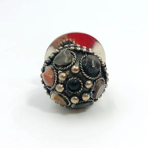 May include: A spherical lapel pin featuring a dark oxidised silver-tone metal base decorated with small silver beads, twisted wire accents, and embedded polished stones in shades of black, grey, brown, and orange, with a flat circular back disc.