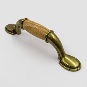 Può includere: Una maniglia per armadio in ottone con un pomello in legno. La maniglia ha una forma curva e un'estremità arrotondata. Il pomello è in legno chiaro.