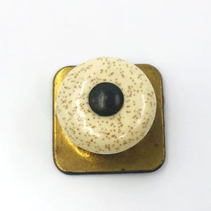 May include: A cabinet knob featuring a round, cream-colored ceramic top with small brown speckles and a dark brown circular center. The knob is mounted on a square, flat, metallic gold-toned base with rounded corners and a smooth, reflective finish.
