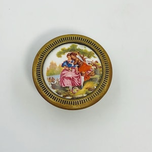 May include: A round, antique brass knob with a painted porcelain insert. The insert depicts a couple in period clothing in a pastoral scene with trees and a lake. The knob has a ridged edge and is likely for a cabinet or drawer.